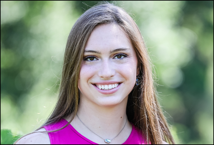 Smiling young woman with brown hair and a pink top with green background outside.