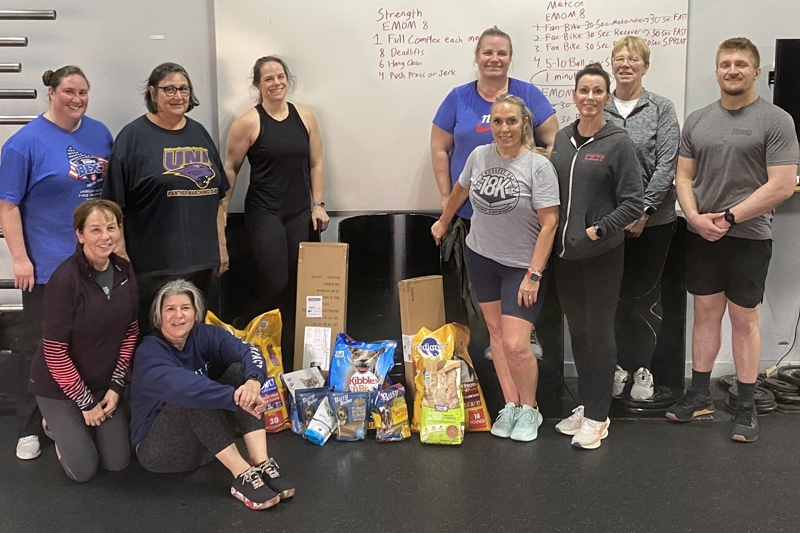 photo of Large group of smiling people in a gym with pet food donations in center.