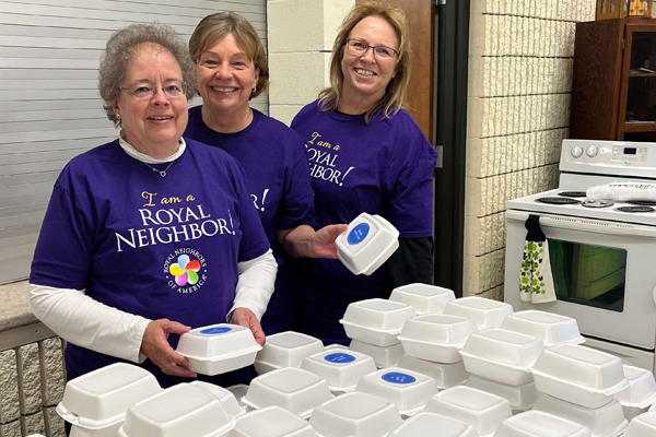 Photo of three happy women wearing purple I am a Royal Neighbor shirts in a kitchen with styrofoam boxes.