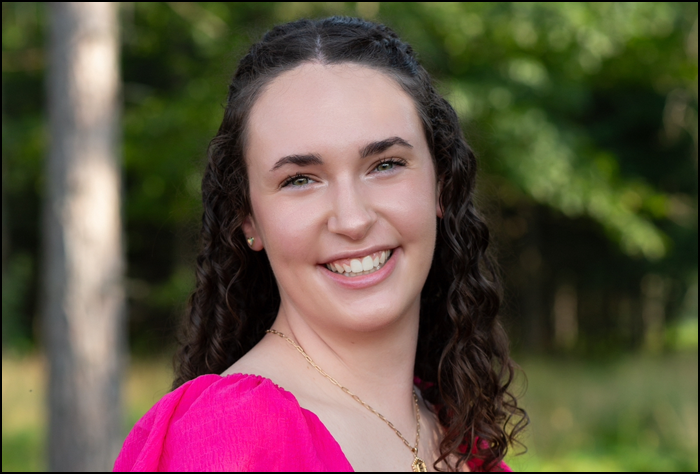Smiling young woman with dark brown curly hair and a pink top with green background outside.