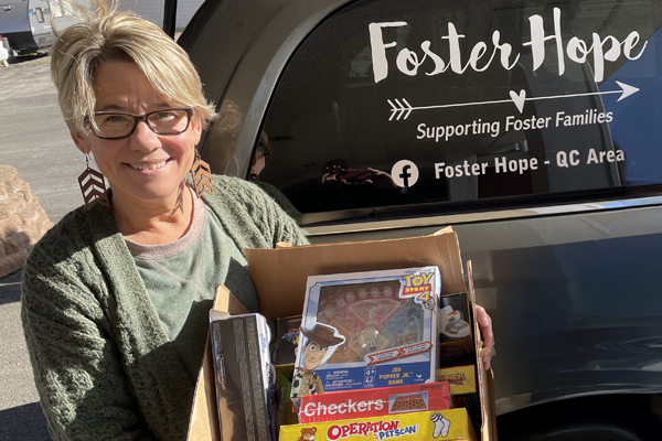 Smiling woman holding a box of toys and games outside of a gray vehicle that says Foster Hope, Supporting Foster Families. Foster Hope - QC Area.