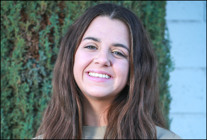 Smiling brunette woman outside in front greenery and white siding.