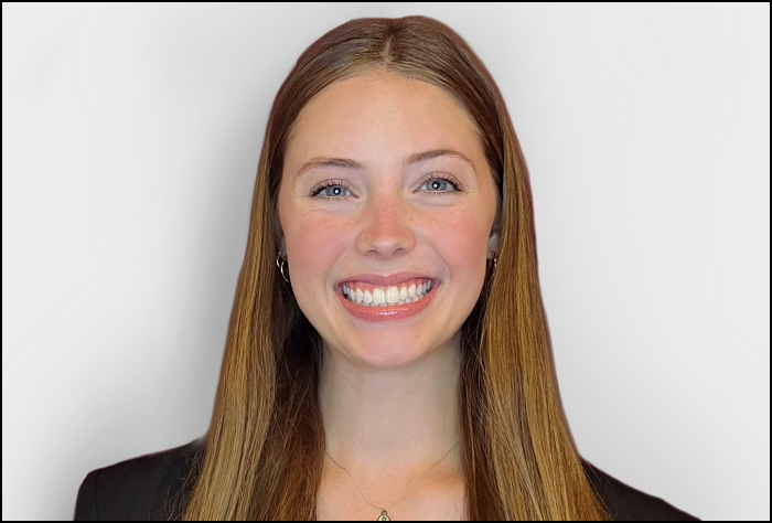 Smiling young woman with long light brown hair and black suite coat with a white background.