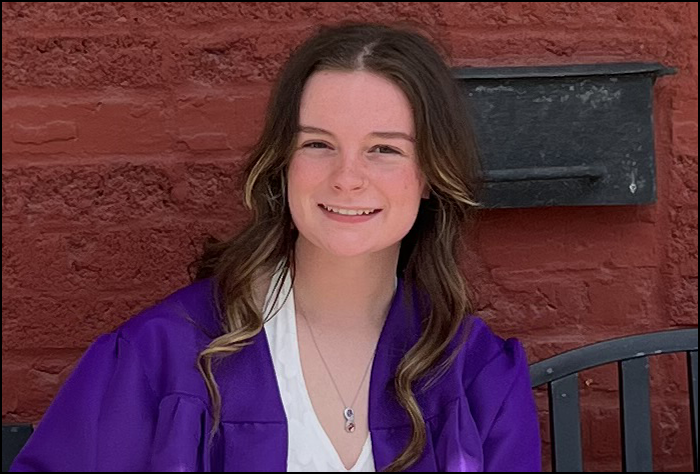 Smiling young woman in a purple graduation gown in front of a brick wall.