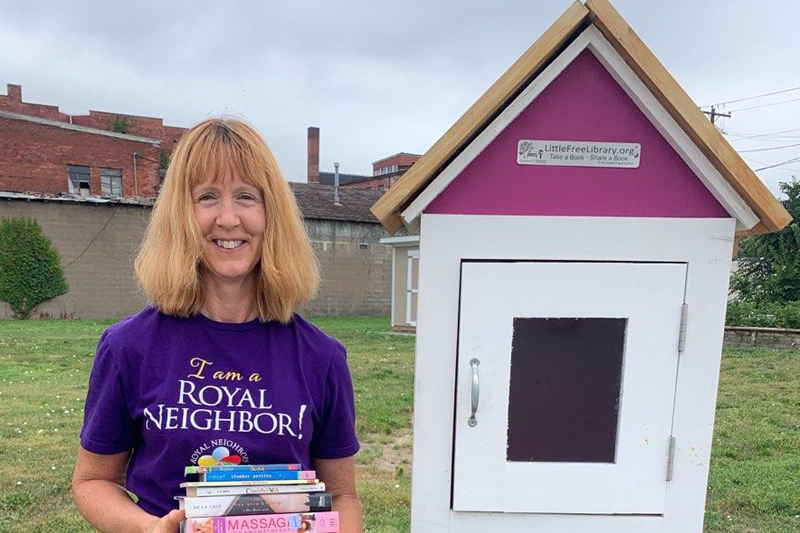 photo of Smiling woman in I am a Royal Neighbors purple t-shirt holding books in front of a Little Free Library box outside.