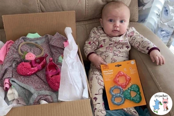 Baby sitting on a tan couch by a box of baby clothing and suppplies.