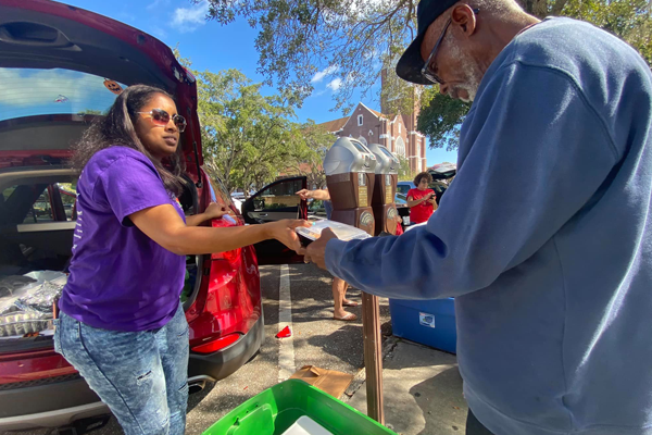 Woman in a purple t-shirt in front of a red car, handing a white food box from a green bin to a gentleman in a blue sweatshirt, glasses, and hat.