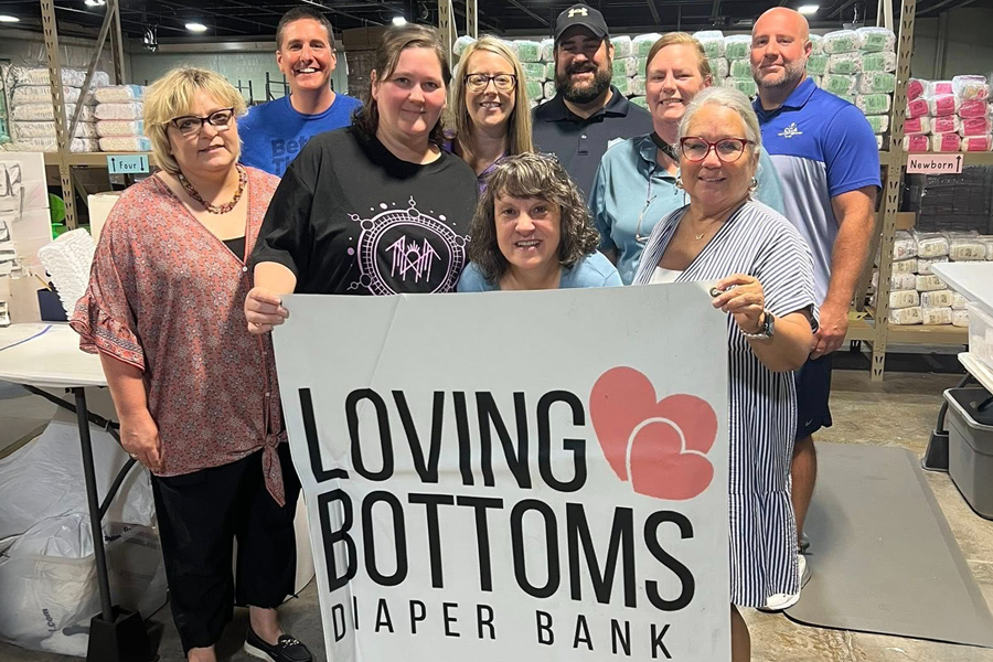 Group of happy people holding a Loving Bottoms Diaper Bank sign in a warehouse of diapers.