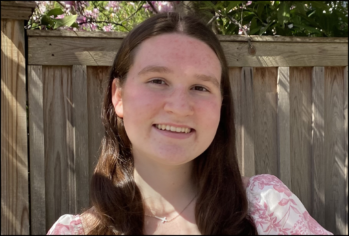 Smiling yougn women outside in front of a fence, with brown hair in a pink and white shirt with sun shining on her.