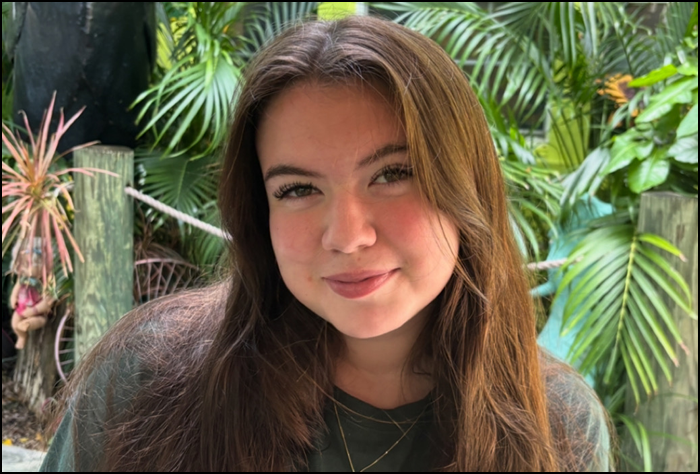 Smiling young woman with brown hair and a gray shirt with a green leaves background.