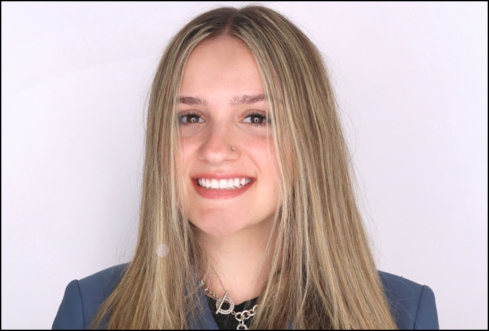 Smiling young blond woman in a gray suite with a lavender background.