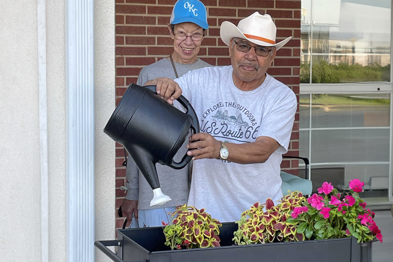 photo of Smiling woman standing behind a man holding a watering can over plants outside in front of a brick building.