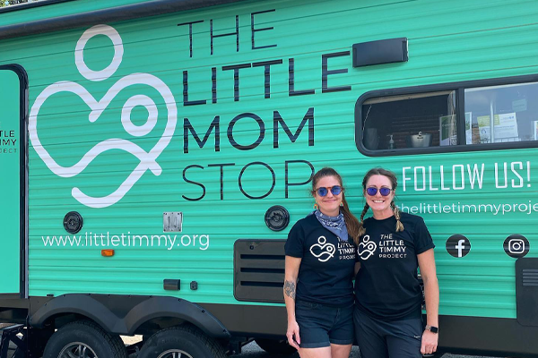 Two smiling women with sunglasses standing in front of a green trailer that says the Little Mom Stop on it.